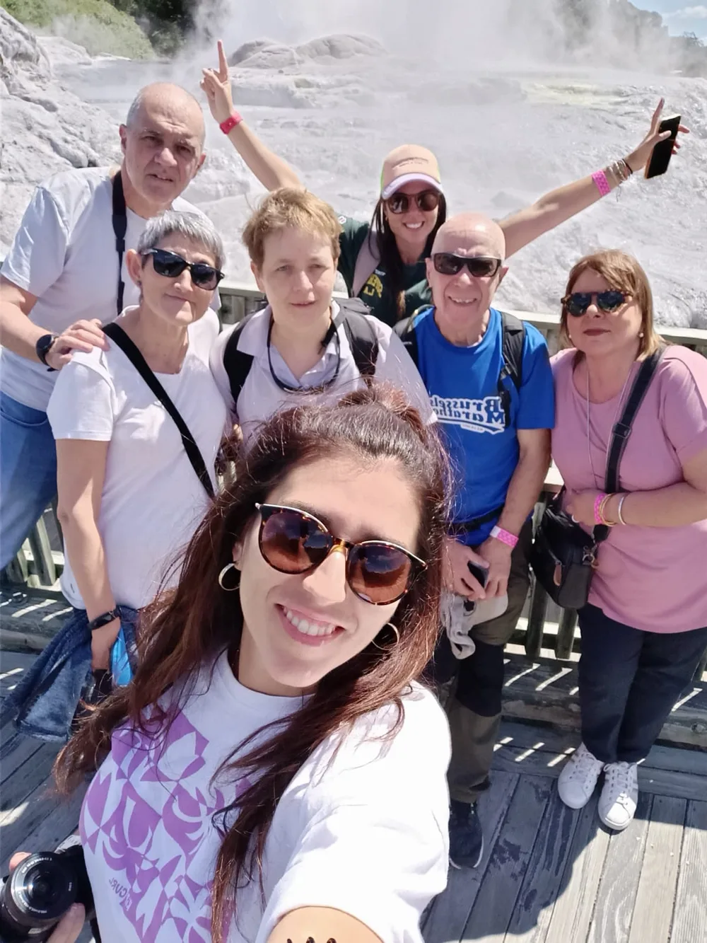 Seven travellers and guides gather on a boardwalk with steam from the Pōhutu Geyser rising behind them.