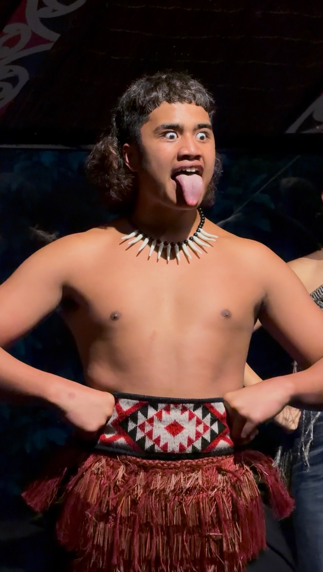 Young Māori man performing a haka at a cultural show, wearing a traditional bone necklace and piupiu skirt, pulling out his tongue in a ceremonial gesture of challenge