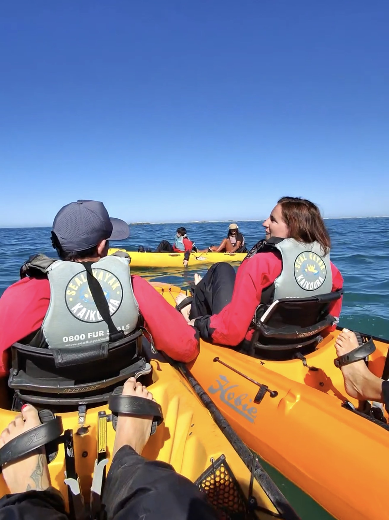 Two travellers sitting in a yellow kayak on the waters of Kaikōura, wearing Seal Kayak Kaikoura life jackets, looking towards another kayak with more travellers under a clear sky