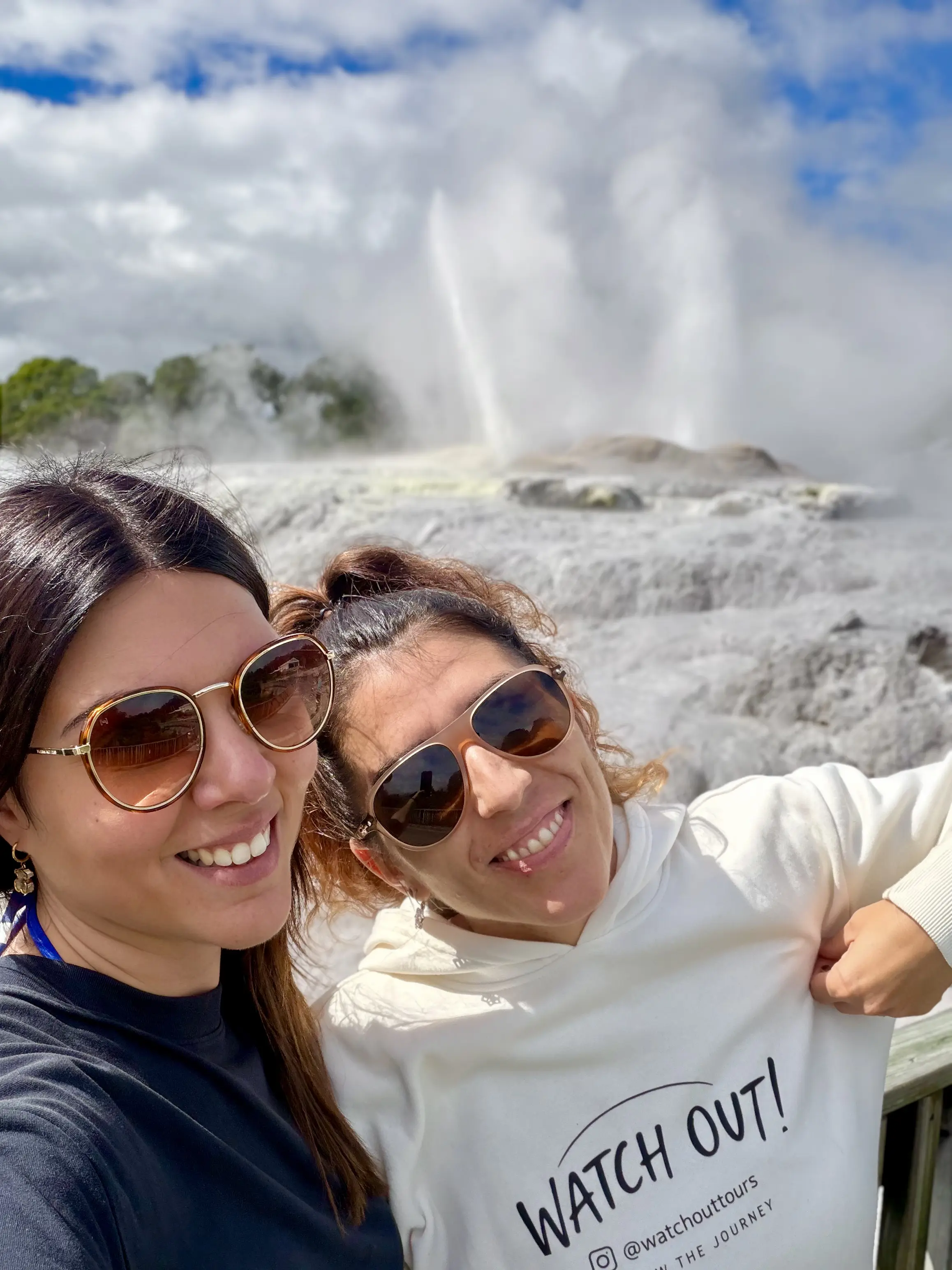 Sylvie and Moni, Watchout Tours guides, smiling in front of a steaming geyser in Rotorua